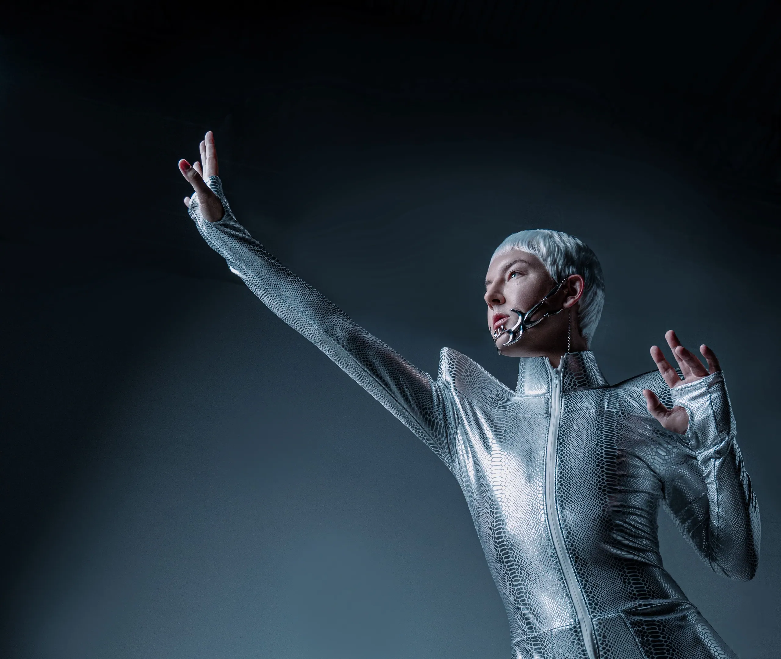 Performer in a silver snakeskin bodysuit reaching upward against a dark blue backdrop